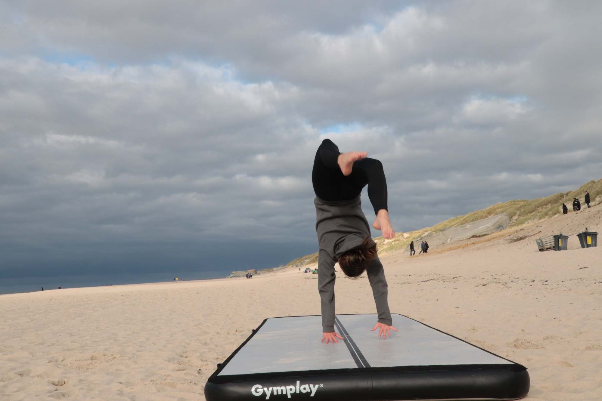 Person laver håndstand på en sort airtrack på en sandstrand under en stormfuld himmel