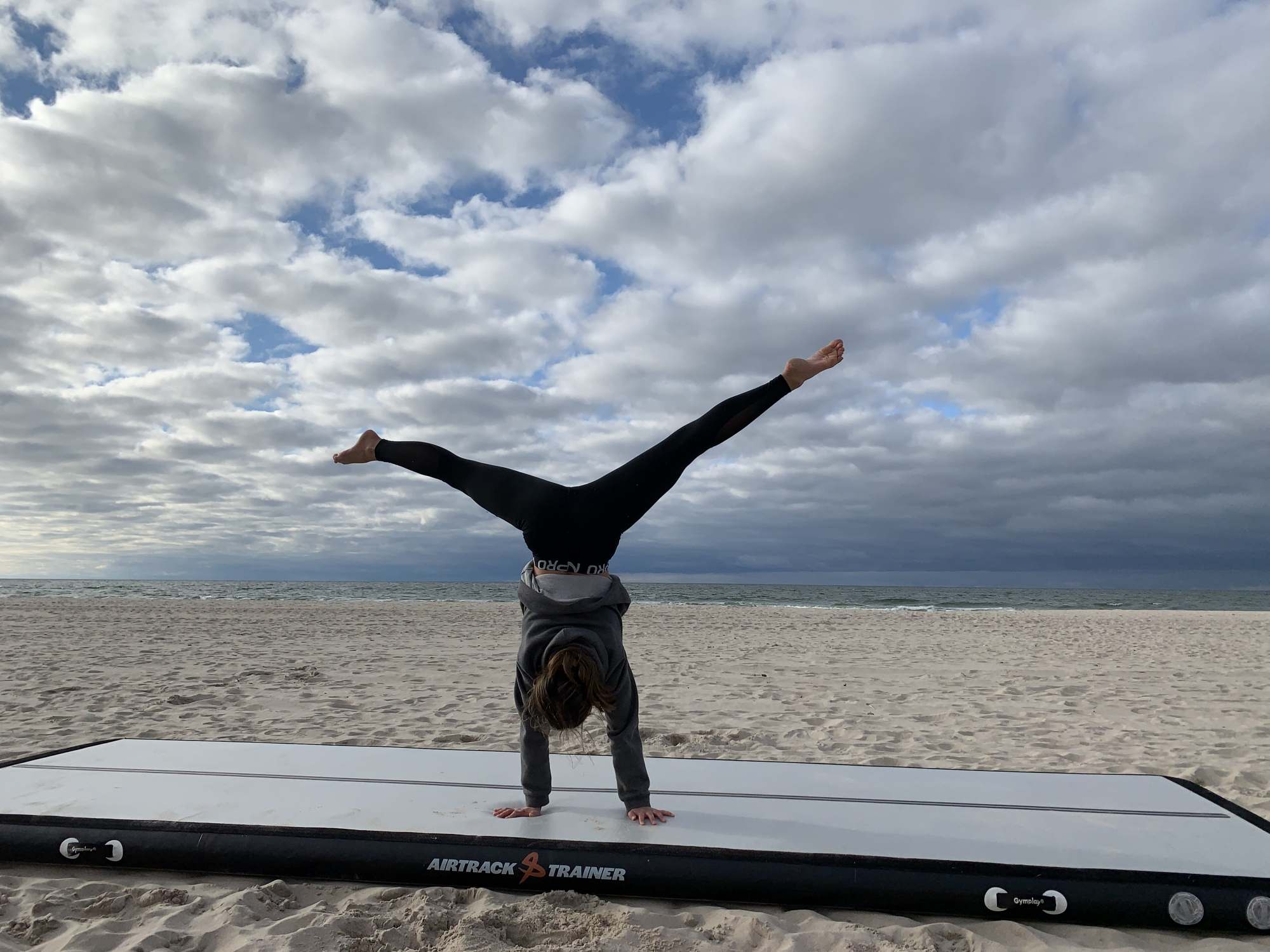 Pige i håndstand med spredte ben på en grå airtrack på en strand under skyet himmel